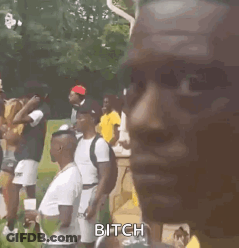 A Man Wearing A Yellow Shirt And A Necklace Is Standing In Front Of A Group Of People . GIF