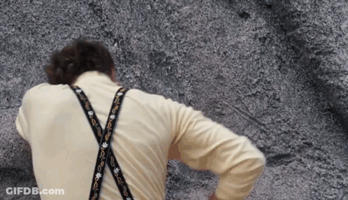 A Man Wearing A Yellow Shirt And Suspenders Is Standing In Front Of A Rock GIF