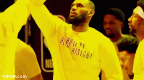 A Man Wearing A Yellow Shirt That Says `` Built By History '' Is Standing In Front Of A Crowd Of People . GIF