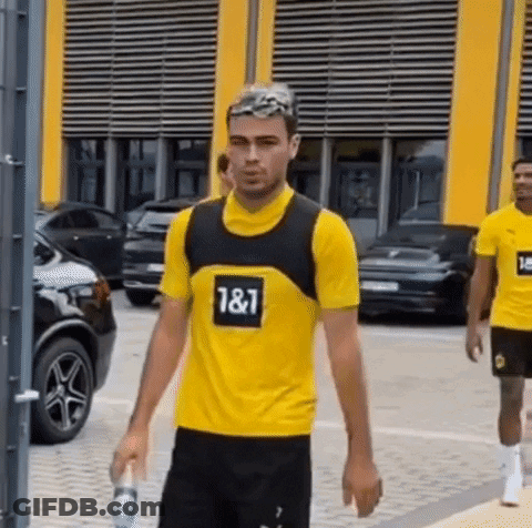 A Man Wearing A Yellow Shirt With The Number 187 On It Is Walking Down A Street Holding A Bottle Of Water . GIF