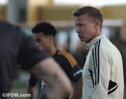 A Man Wearing An Adidas Jacket Stands In Front Of A Group Of Men GIF