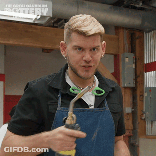 A Man Wearing An Apron Is Holding A Pair Of Scissors In Front Of A Sign That Says The Great Canadian Pottery GIF