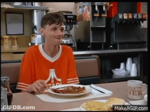 A Man Wearing An Atari Shirt Is Sitting At A Table With A Plate Of Food On It GIF
