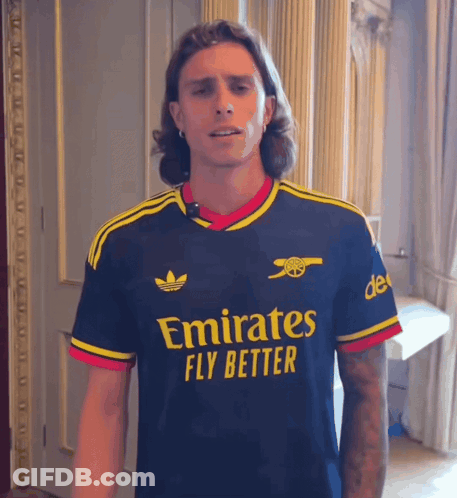 A Man Wearing An Emirates Fly Better Shirt Stands In A Room GIF
