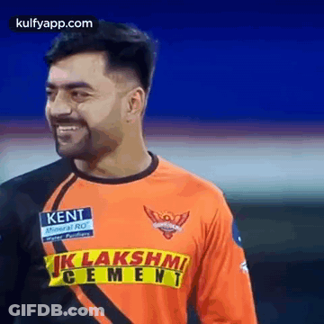 A Man Wearing An Orange And Black Jersey With A Yellow Stripe On The Front Is Smiling . GIF