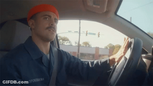A Man Wearing An Orange Beanie And A Blue Shirt With The Name Opokukwa On It Is Driving A Car GIF