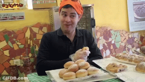 A Man Wearing An Orange Hat Is Sitting At A Table With A Bunch Of Donuts GIF