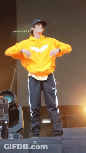A Man Wearing An Orange Hoodie And Black Pants Is Dancing On A Stage GIF
