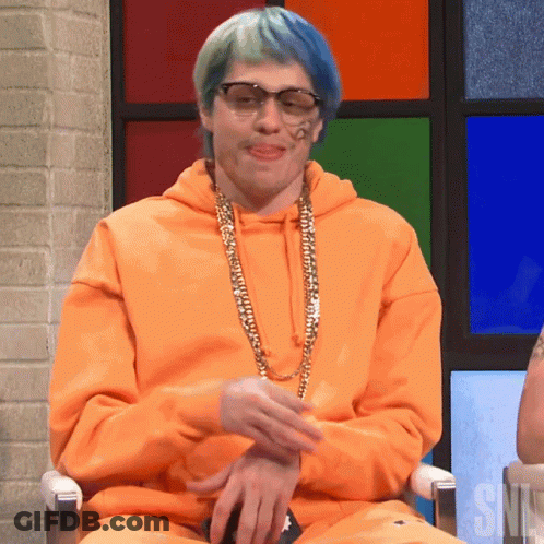 A Man Wearing An Orange Hoodie And Sunglasses Is Sitting In Front Of A Snl Sign GIF