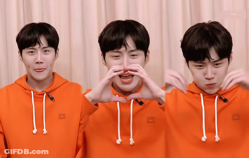 A Man Wearing An Orange Hoodie Is Making A Heart With His Hands GIF