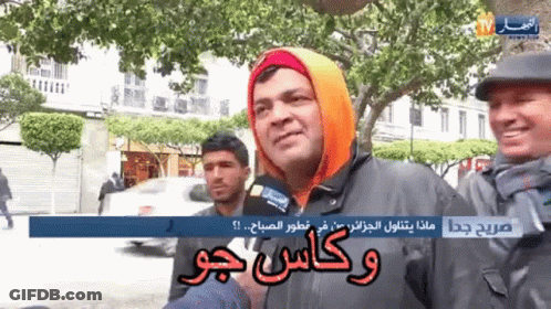 A Man Wearing An Orange Hoodie Is Talking Into A Microphone While Another Man Stands Behind Him GIF
