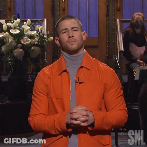 A Man Wearing An Orange Jacket And A Grey Turtleneck Is Standing In Front Of A Snl Sign GIF