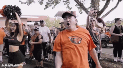 A Man Wearing An Orange Shirt With A Buffalo Bills Logo Stands In Front Of A Cheerleader GIF