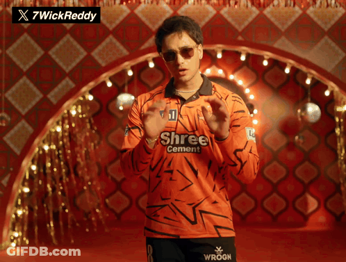 A Man Wearing An Orange Shree Cement Shirt Stands In Front Of A Checkered Wall GIF