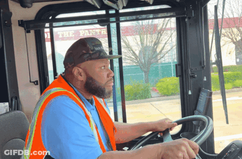 A Man Wearing An Orange Vest And A Hat Is Driving A Bus GIF