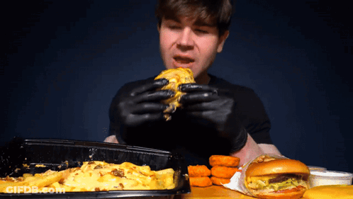 A Man Wearing Black Gloves Eats A Cheeseburger GIF