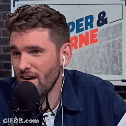 A Man Wearing Ear Buds Is Speaking Into A Microphone In Front Of A Sign That Says Der & Rne GIF
