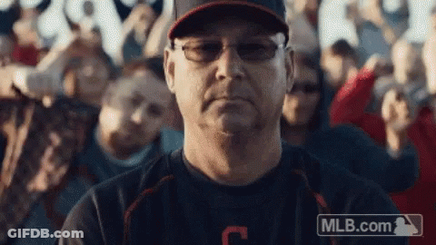 A Man Wearing Glasses And A Baseball Cap Is Sitting In Front Of A Crowd Of People . GIF