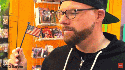 A Man Wearing Glasses And A Black Hat Holds An American Flag GIF