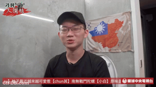 A Man Wearing Glasses And A Black Hat Is Sitting In Front Of A Wall With A Map Of China On It GIF