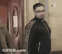 A Man Wearing Glasses And A Black Jacket Is Standing In Front Of A Red Door . GIF