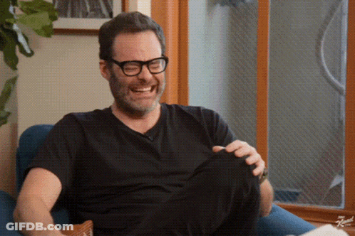 A Man Wearing Glasses And A Black Shirt Is Sitting In A Chair Laughing GIF