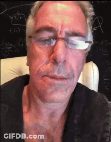 A Man Wearing Glasses And A Black Shirt Is Sitting In Front Of A Blackboard GIF