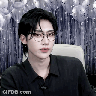 A Man Wearing Glasses And A Black Shirt Is Sitting In Front Of Balloons GIF