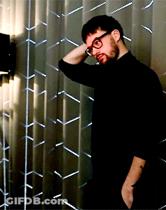 A Man Wearing Glasses And A Black Shirt Is Standing In Front Of A Metal Wall GIF