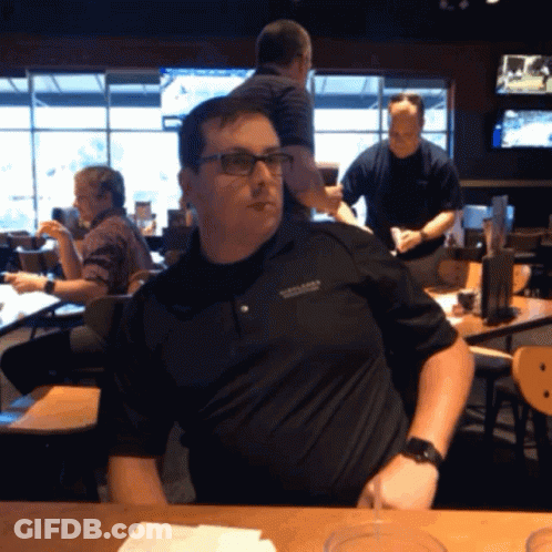 A Man Wearing Glasses And A Black Shirt That Says ' Oklahoma ' On It Sits At A Table In A Restaurant GIF