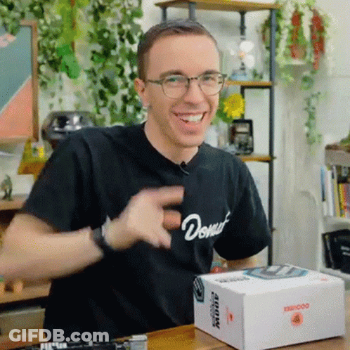 A Man Wearing Glasses And A Black Shirt With The Word Donut On It Is Sitting At A Table With A Box On It . GIF