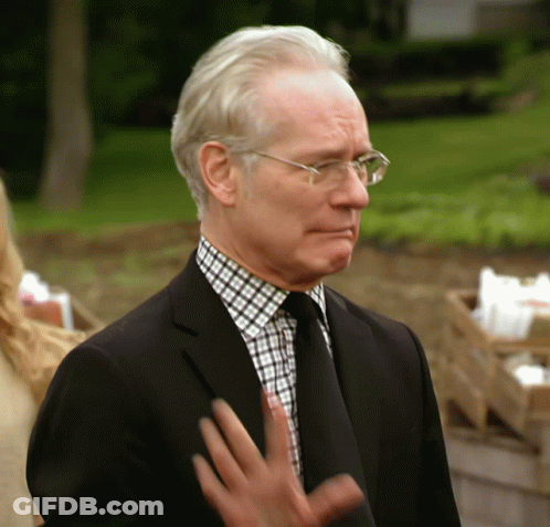 A Man Wearing Glasses And A Black Suit Stands In Front Of A Green Field GIF