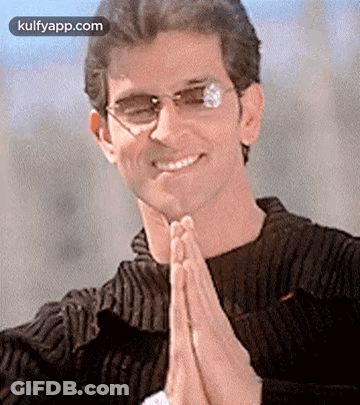 A Man Wearing Glasses And A Black Sweater Is Smiling And Praying With His Hands Folded . GIF