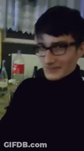 A Man Wearing Glasses And A Black Turtleneck Is Smiling In Front Of A Coke Bottle GIF