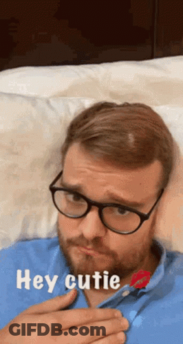 A Man Wearing Glasses And A Blue Shirt Is Laying On A Bed With The Words Hey Cutie Above Him GIF