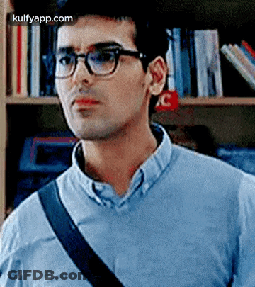 A Man Wearing Glasses And A Blue Shirt Is Standing In Front Of A Bookshelf In A Library . GIF