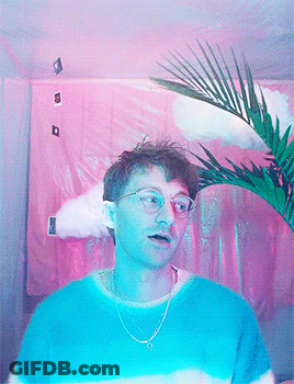 A Man Wearing Glasses And A Blue Shirt Stands In Front Of A Pink Curtain GIF