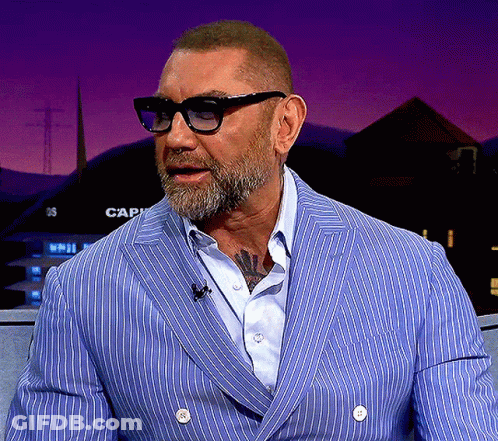 A Man Wearing Glasses And A Blue Striped Jacket Is Sitting In Front Of A Sign That Says Cap GIF