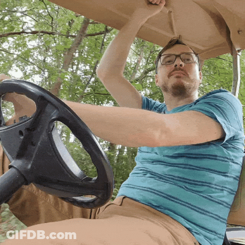 A Man Wearing Glasses And A Blue Striped Shirt Is Driving A Golf Cart GIF