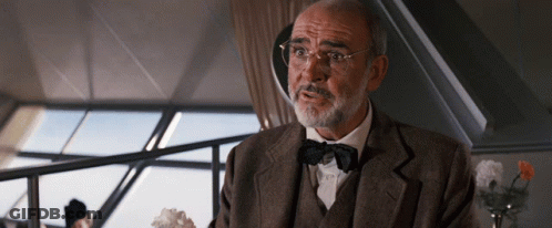 A Man Wearing Glasses And A Bow Tie Is Sitting In Front Of A Window GIF