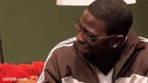 A Man Wearing Glasses And A Brown Jacket Is Sitting On A Red Couch . GIF