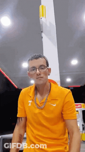 A Man Wearing Glasses And A Chain Is Standing In Front Of A Gas Pump . GIF