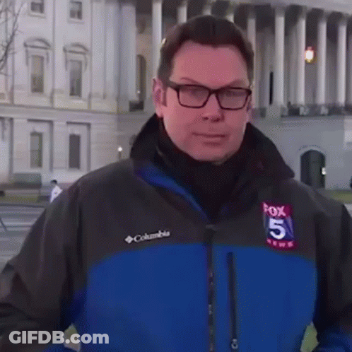 A Man Wearing Glasses And A Columbia Jacket Is Standing In Front Of A Large Building . GIF