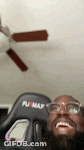 A Man Wearing Glasses And A Furmax Gaming Chair Is Smiling In Front Of A Ceiling Fan . GIF