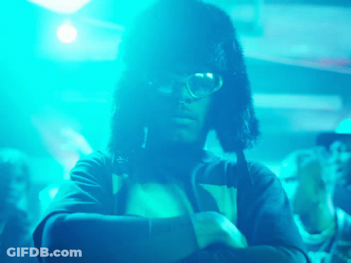 A Man Wearing Glasses And A Furry Hat Is Standing In Front Of A Crowd Of People GIF