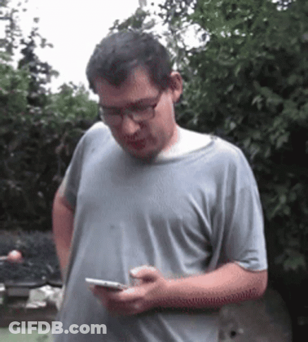 A Man Wearing Glasses And A Gray Shirt Is Looking At His Phone GIF