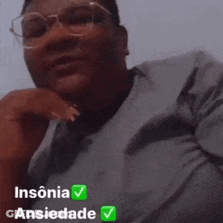 A Man Wearing Glasses And A Gray Shirt Is Sitting Down With His Hand On His Chin And Looking At The Camera . GIF