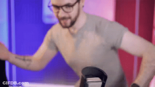 A Man Wearing Glasses And A Gray Shirt Is Standing In Front Of A Microphone . GIF