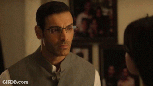 A Man Wearing Glasses And A Gray Vest Looks At Another Man GIF