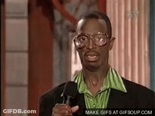A Man Wearing Glasses And A Green Shirt Is Pointing At The Camera While Talking Into A Microphone . GIF
